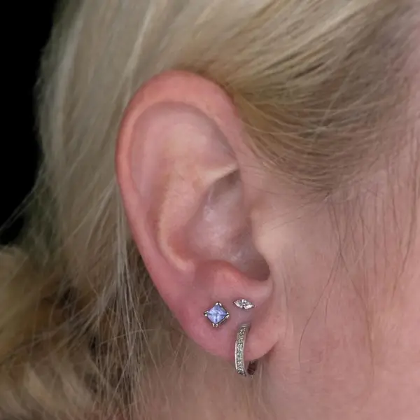 Stacked lobe piercings with a blue princess-cut gem, a marquise stud, and a pavé huggie hoop by T (Piercing Piranha) at Modified Tattoo Gallery in Winter Park, FL